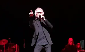 A male singer in a black suit and glasses points to the sky while holding a microphone on a dark stage.