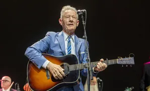 A man in a blue suit plays an acoustic guitar on stage, with a microphone in front of him.