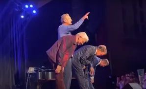 Four men in suits bow on stage, with one man in a blue shirt standing behind them pointing.