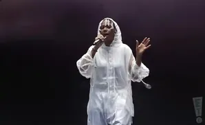 A performer in a white hooded outfit sings into a microphone, with a black background.