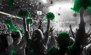 A crowd cheers with green pom-poms and confetti as green banners fly, with a bright light in the background.