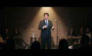 Back view of a man in a suit speaking to an audience under spotlights.