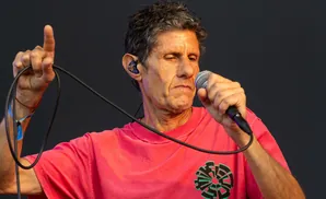 A man with grey hair sings into a microphone, wearing a pink shirt and an ear monitor.
