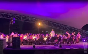 An orchestra performs under purple lights on an outdoor stage, with a conductor in a white suit.