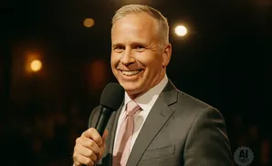 A man in a suit and tie smiles and holds a microphone.