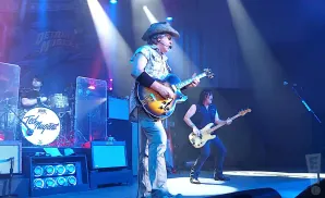 Ted Nugent performs on stage with his band, playing guitar and wearing a cowboy hat.