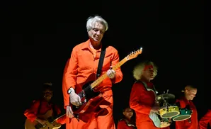 Musicians in orange jumpsuits perform on stage with David Byrne in the foreground playing a red guitar.