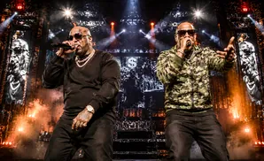 Two rappers in chains and sunglasses perform on stage with a large screen behind them showing their faces and dollar signs.