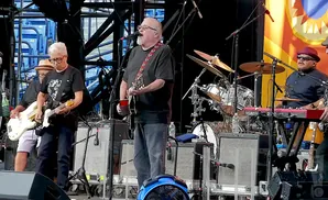 A band performs on stage. A guitarist in glasses wears a black t-shirt and jeans, playing a white electric guitar.