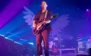 A guitarist plays on a stage with large white wings projected behind him, bathed in purple and blue lights.