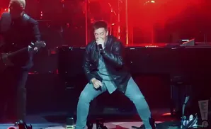A male singer in a leather jacket and grey pants sings into a microphone on stage.