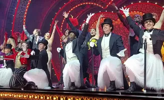 A cast of performers in elaborate costumes bow and wave from a stage.