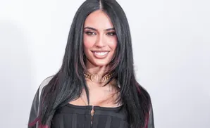 A woman with long dark hair with pink streaks smiles, wearing a black ribbed top and a gold 'CD' choker.