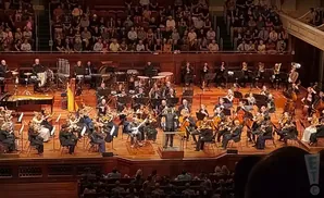 An orchestra performs on stage with a conductor in front, and an audience in the background.