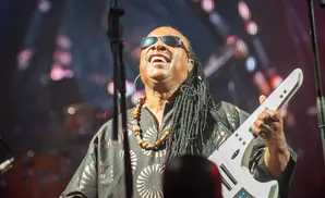 Stevie Wonder performs on a white keytar, wearing sunglasses and a patterned shirt with a beaded necklace.