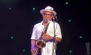 A Black man in a white hat and patterned shirt plays a saxophone on stage with green lights in the background.