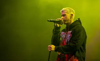 A singer with bleached hair performs on stage, wearing a black hoodie with a red graphic.