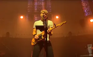Man in a striped shirt plays a yellow electric guitar on stage with warm lighting.