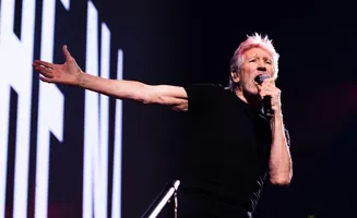 Roger Waters performs on stage with his arm outstretched and holding a microphone.