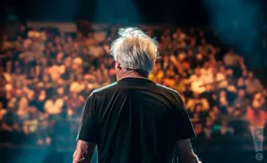Man with white hair facing away from camera on stage, with blurred audience in background.