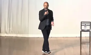 A bald man in a suit speaks into a microphone, while standing on a wooden stage.
