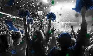 A crowd cheers at a concert, with confetti falling and people holding up blue pompoms.
