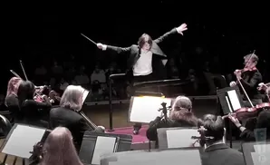 An orchestra conductor directs musicians in a dark theater.