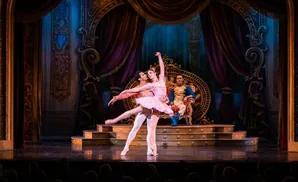 A ballet couple dances on stage in front of a prince sitting on a throne, with an orchestra playing in the background.