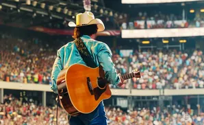 A man in a cowboy hat plays an acoustic guitar on stage in front of a large, blurred audience.