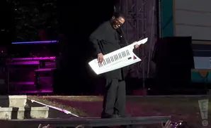 A man in a black outfit plays a white keytar on a stage with purple lighting.