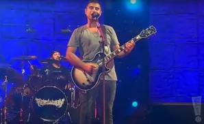 A male guitarist sings and plays a guitar on stage with a drummer in the background.