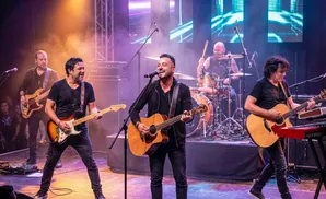 A rock band performs on stage, with lead singer playing acoustic guitar, a guitarist, bassist, keyboardist, and drummer.