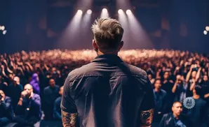 Man with tattoos on arms faces a large cheering crowd under stage lights.