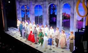 A cast of actors in historical costumes stand on a stage in front of an elaborate set depicting a garden with arched windows.