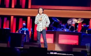 A man in a plaid shirt stands center stage with a microphone, while a drummer plays in the background.