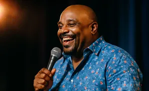 A bald Black man with a goatee laughs heartily while holding a microphone, wearing a blue shirt with a floral pattern.