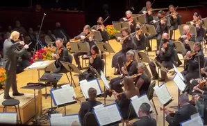 An orchestra conductor directs musicians playing violins and other instruments on stage.