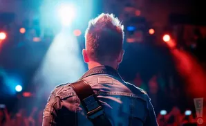 Back view of a musician with spiky hair on stage, wearing a denim jacket with a guitar strap, lit by stage lights.