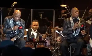 Three Black men in sparkly suits perform on stage. Two sing into microphones, one plays drums.