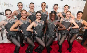 Dancers in sparkly black outfits pose on a red carpet in front of a 