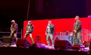 Four men on stage wearing sparkling jackets and hats, performing in front of a red backdrop.