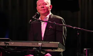 Man playing keyboard with pink lighting, wearing a suit with a polka dot pocket square.