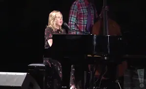 A blonde woman sings at a piano, with a man playing the double bass behind her.