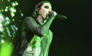 A heavily tattooed singer with green lighting and makeup sings into a microphone.