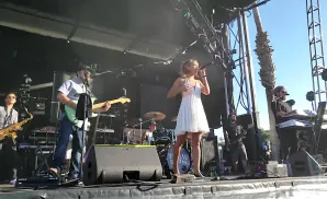 A band performs on an outdoor stage, with a female singer in a white dress, a guitarist, a saxophonist, and a keyboardist.