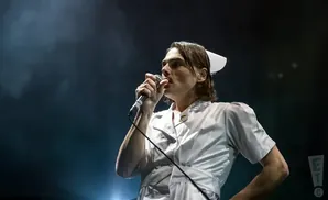A singer in a nurse's uniform and cap performs into a microphone on a dimly lit stage.