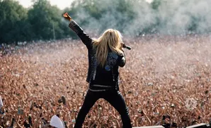 Singer with long blonde hair in a black studded jacket and pants, singing into a microphone with a large crowd cheering.