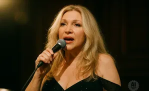 Woman singing into a microphone, with blonde hair and wearing a black dress.