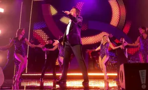 Singer in a black suit on stage with backup dancers in sequined outfits.