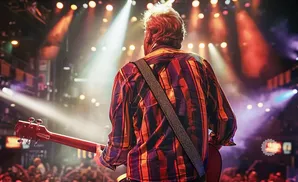 Musician playing guitar on stage with bright lights and a cheering crowd.